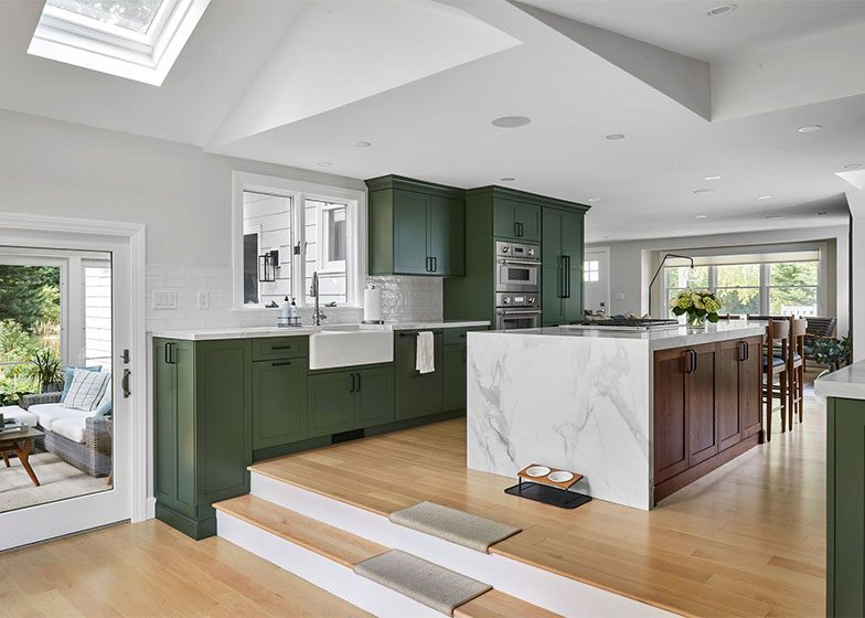 Modern kitchen with green cabinets, a marble island, and stainless steel appliances. Hardwood floors lead to a bright, open living area. A cat ramp is near the steps.