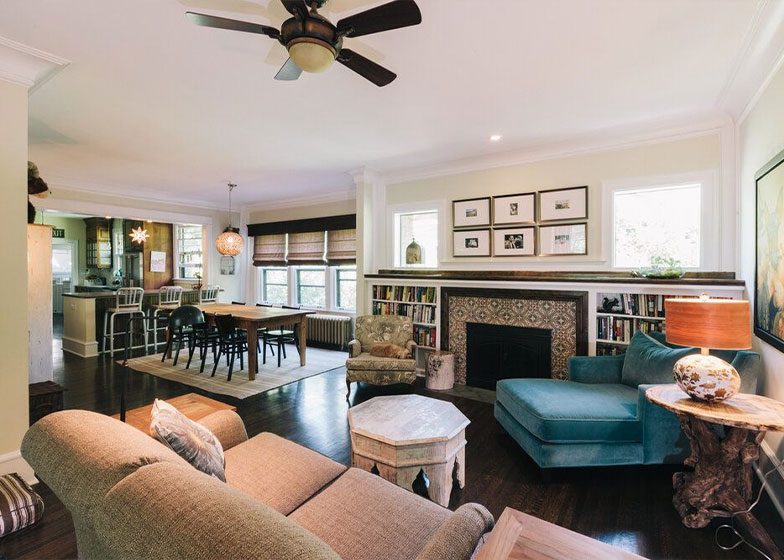 Cozy living room with a fireplace, sofa, chairs, and bookshelves. Open layout connects to dining area and kitchen. Ceiling fan and framed pictures add decor.